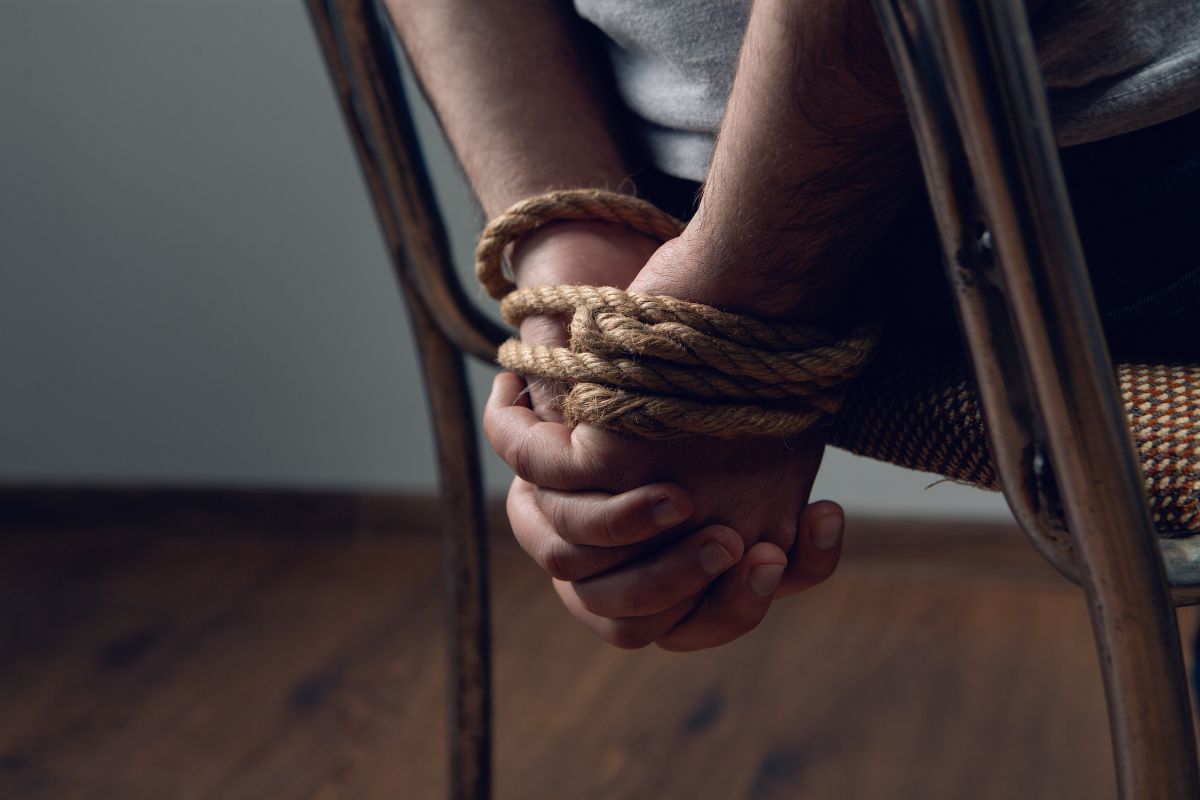 Person’s hands tied with rope behind a chair. Person’s hands tied with rope behind a chair.