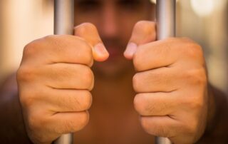 A man's hands are gripping jail bars tightly.