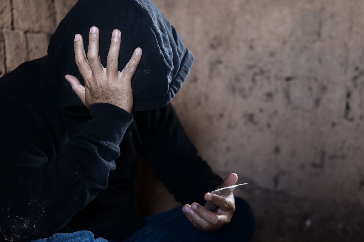 A man is covering their face while holding a small packet of drugs.