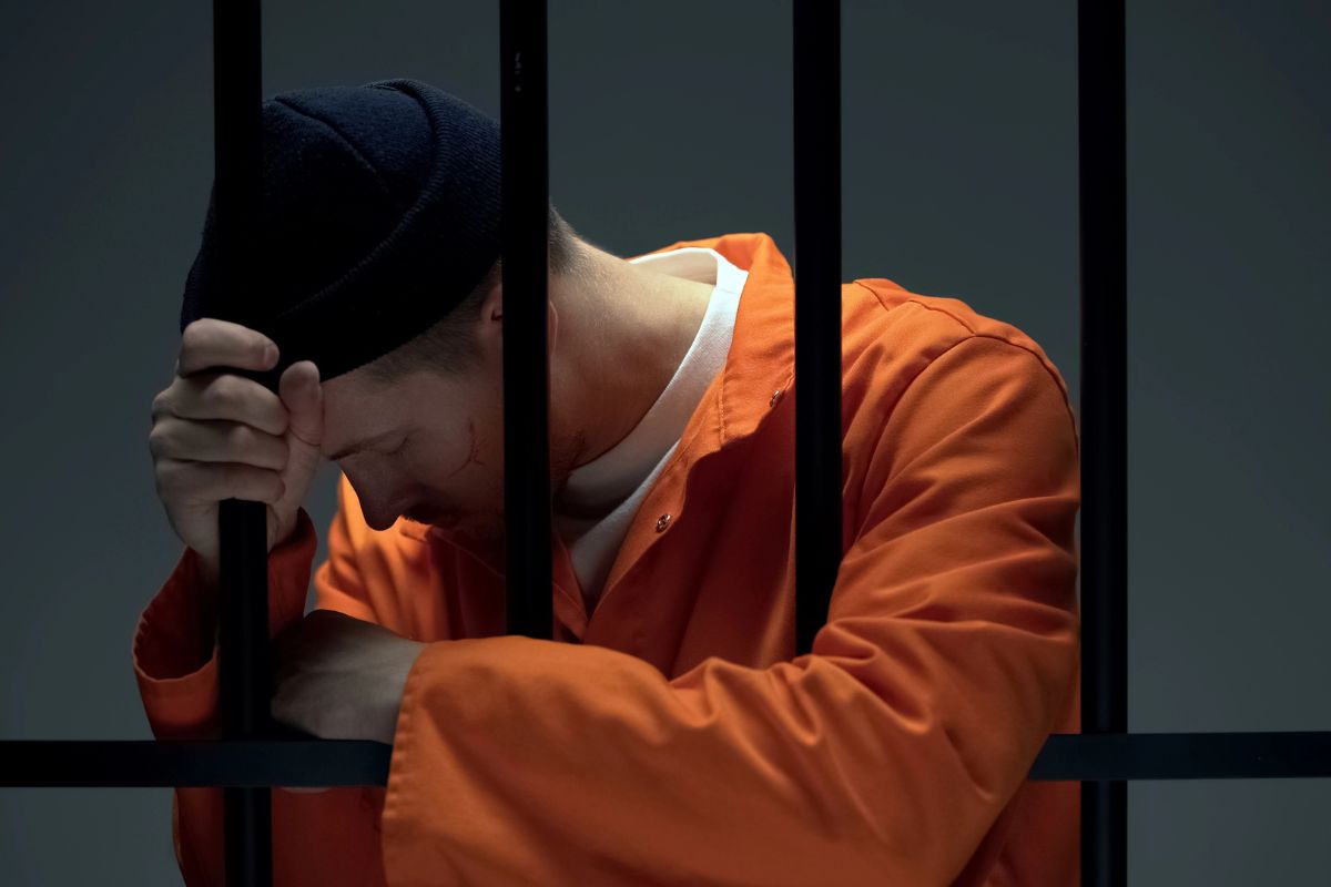 A man in a prison uniform behind bars with his head bowed.