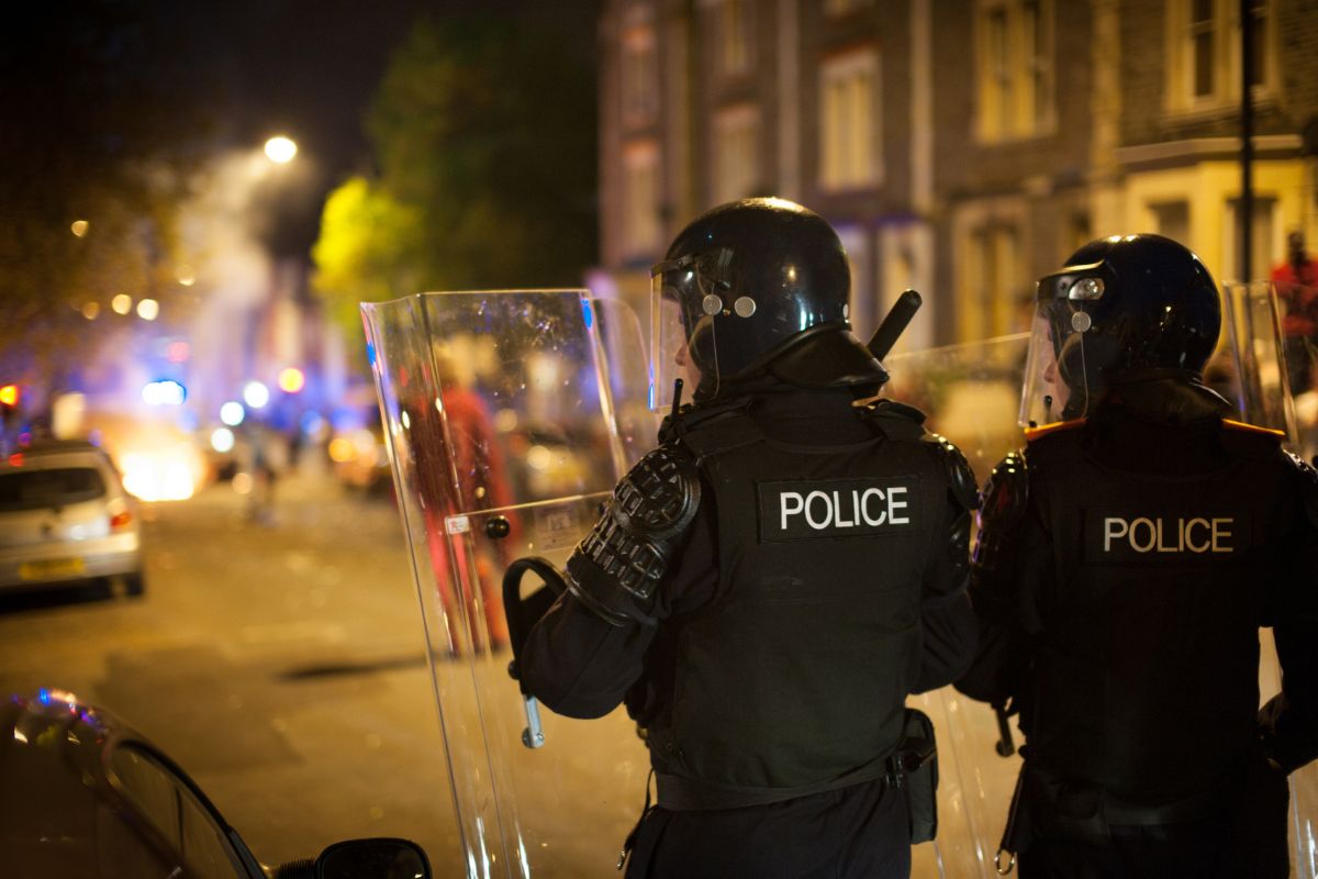 Police officers in full riot gear with shields raised.