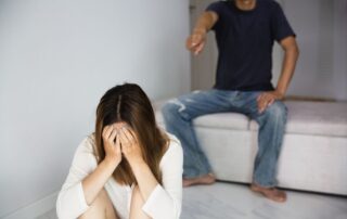 A young woman covering her face while a man points at her in a home.