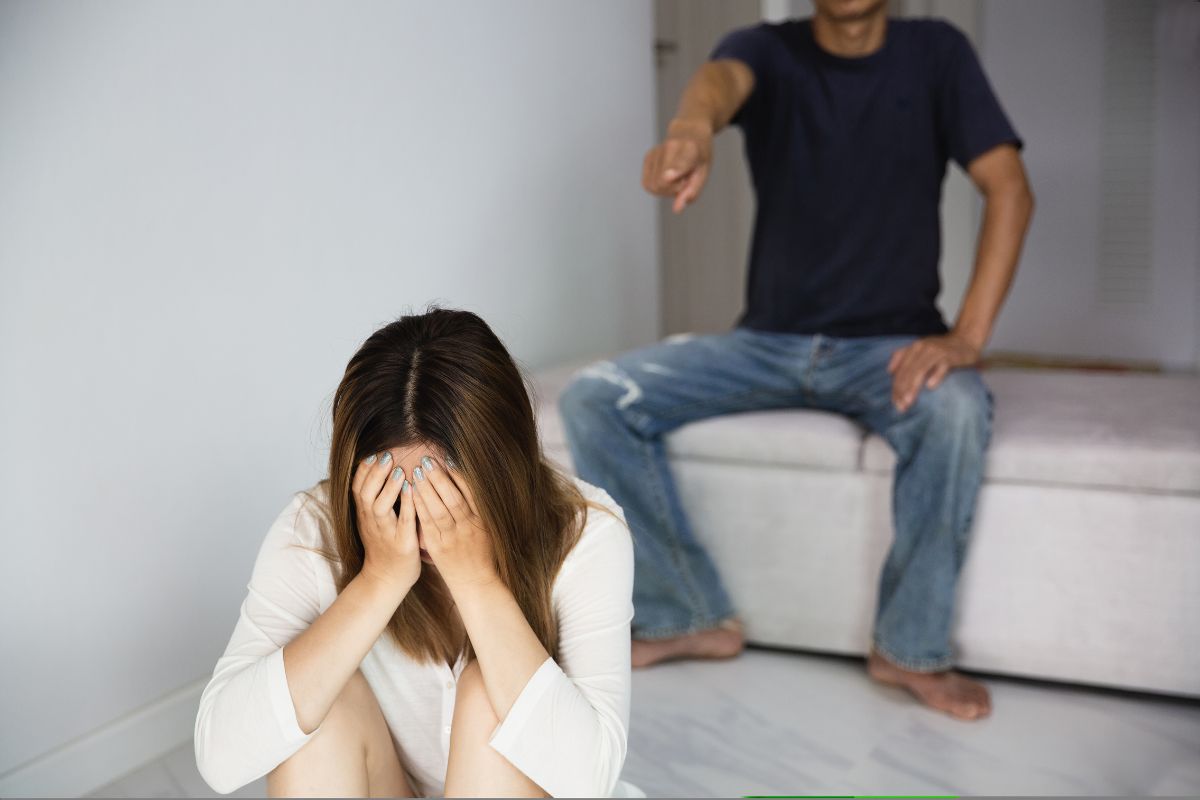A young woman covering her face while a man points at her in a home.