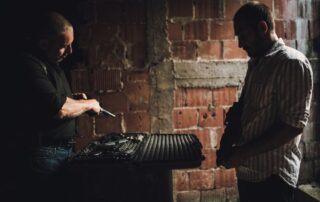 Two men in a dark room exchanging firearms from an open case.