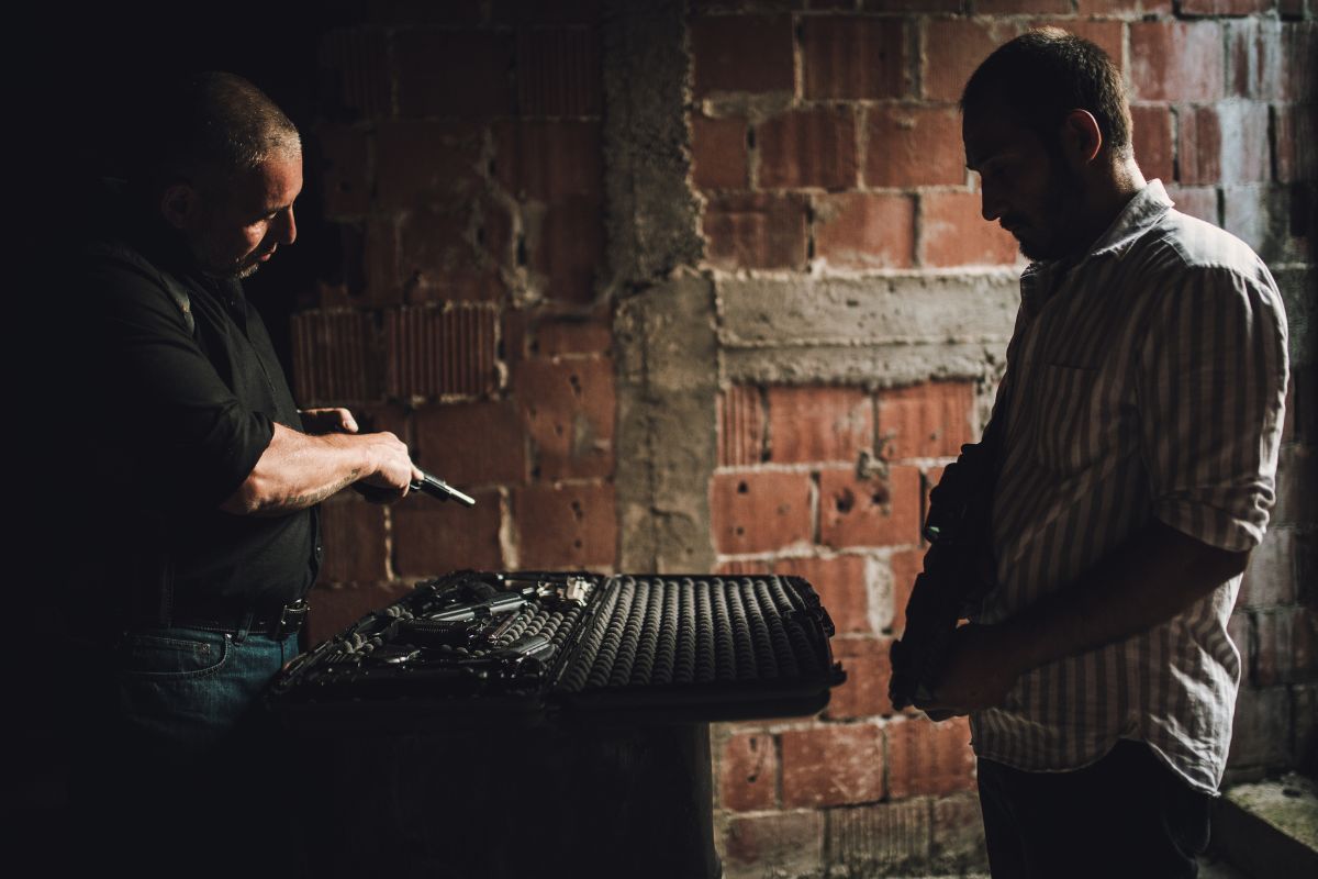 Two men in a dark room exchanging firearms from an open case.