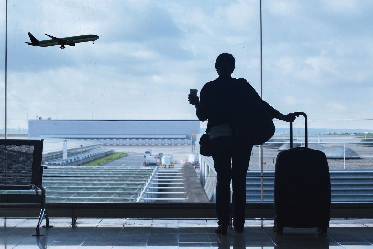 A traveler with luggage looking out at a plane taking off through an airport window.