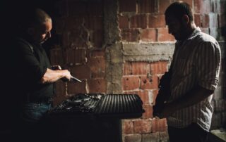 Two men in a dark room exchanging firearms from an open case.
