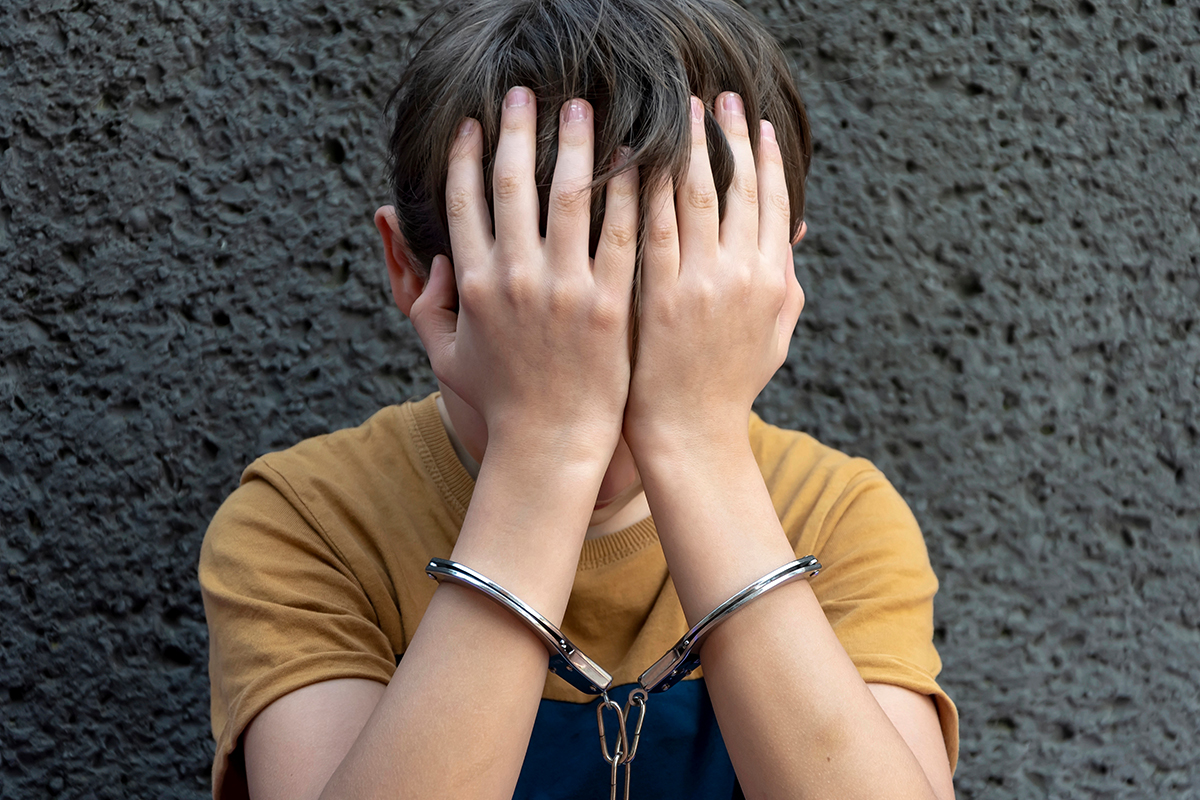 A young boy in hand cuffs with his hands covering his face