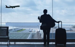 A traveler with luggage looking out at a plane taking off through an airport window.