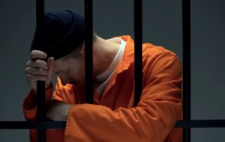 A man in a prison uniform behind bars with his head bowed.